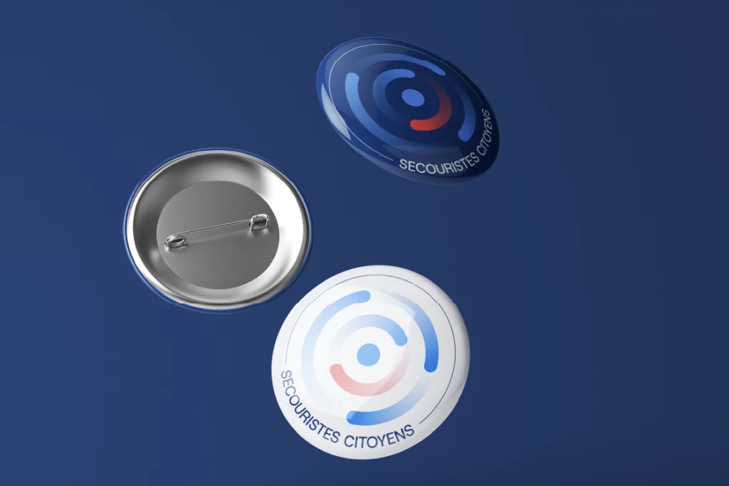 Trois badges métalliques arborant le logo des Secouristes Citoyens en bleu et en blanc.
